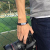 Riptide microfibre leather and stainless steel medical alert bracelet worn by a a male model in front of a sports field, holding a camera.